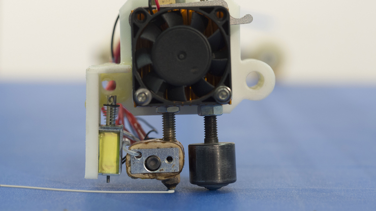 a close-up picture of the head of the device. You see from left to right a scraper to remove contents, an extruder which prints lines and a ball caster that prevents the extruder from hitting the board. On top of that is a large ventilator to cool the hea